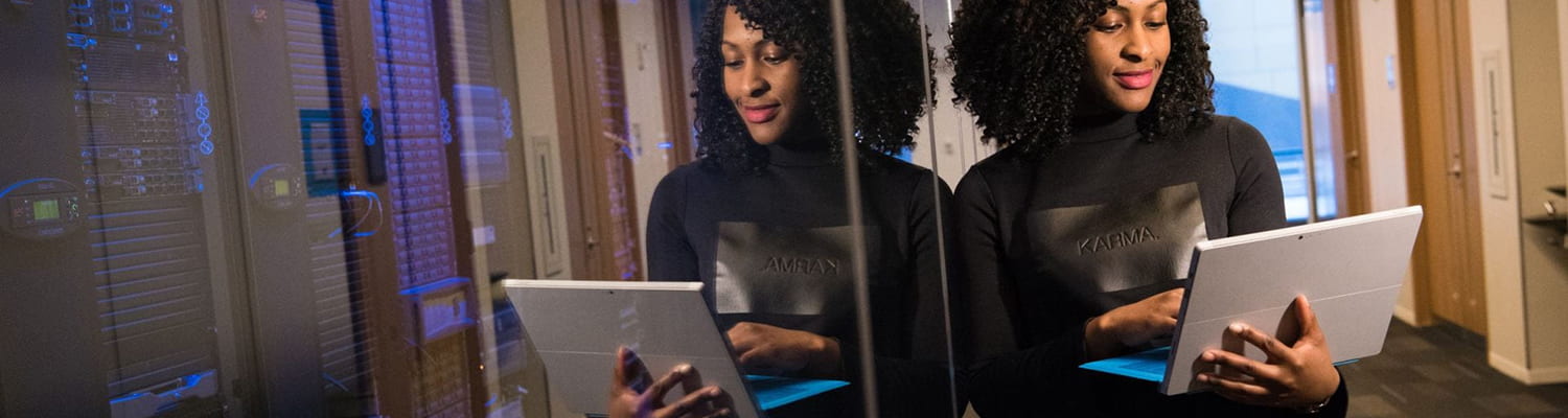 Image: A young, black woman near a computer room. She is holding a laptop and wearing a shirt that has a patch that says 'karma'. 